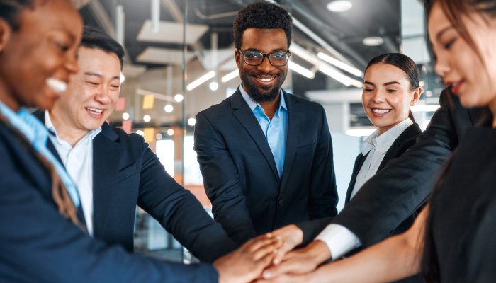 A team of successful businessmen of different nationalities in suits celebrating victory, success while standing in a modern office with their hands clasped together.